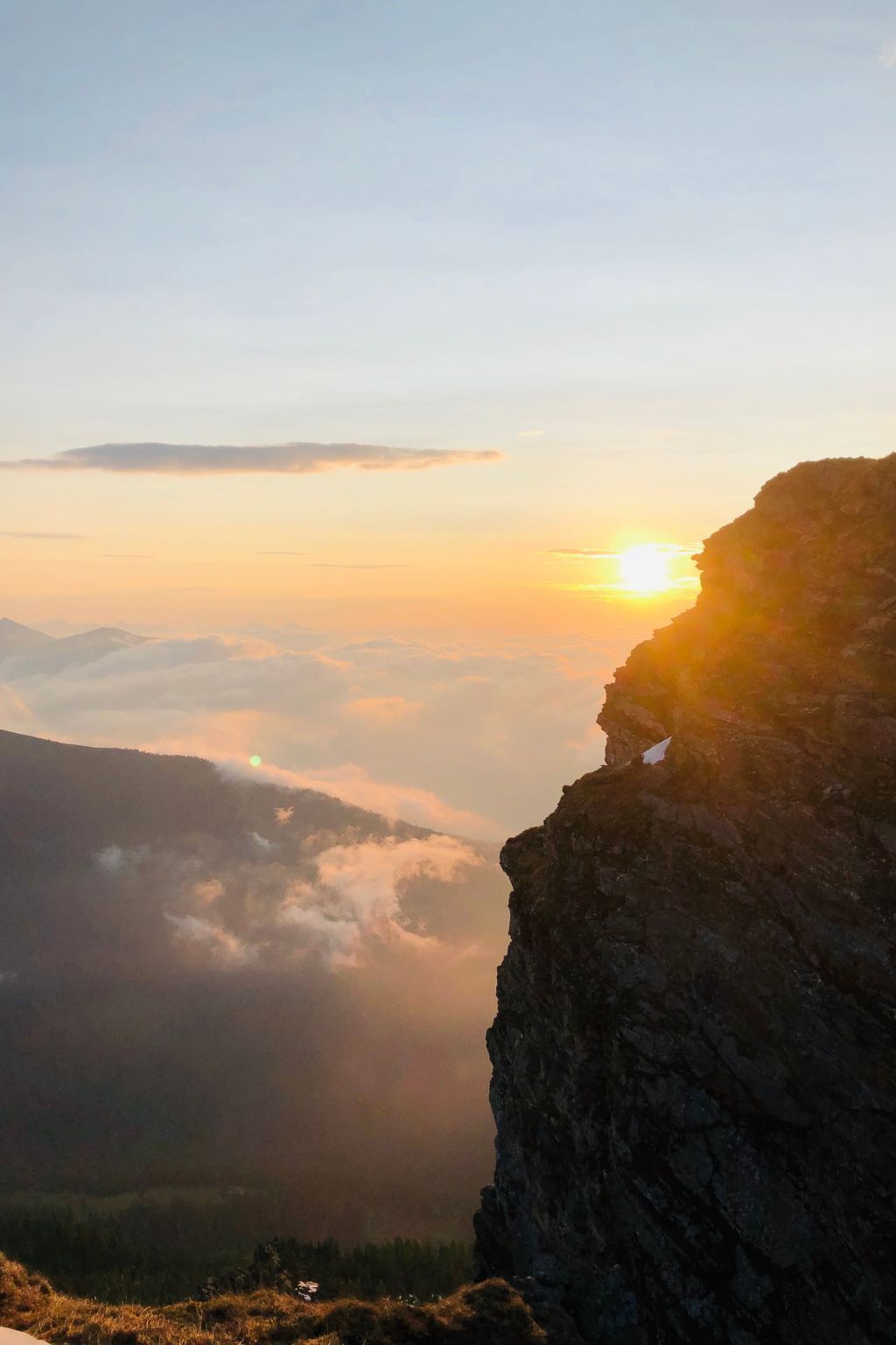 Paisatge de muntanya amb sol ixent o ponent, núvols baixos i un penya-segat rocós.