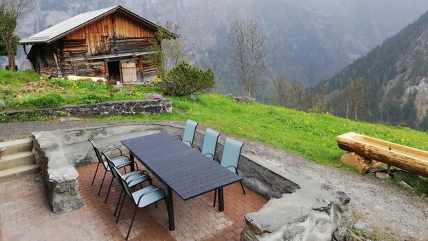 A rustic wooden cabin with a stone patio and outdoor seating area, surrounded by lush greenery and mountains. - Husmättli by Interhome (Lauterbrunnen)