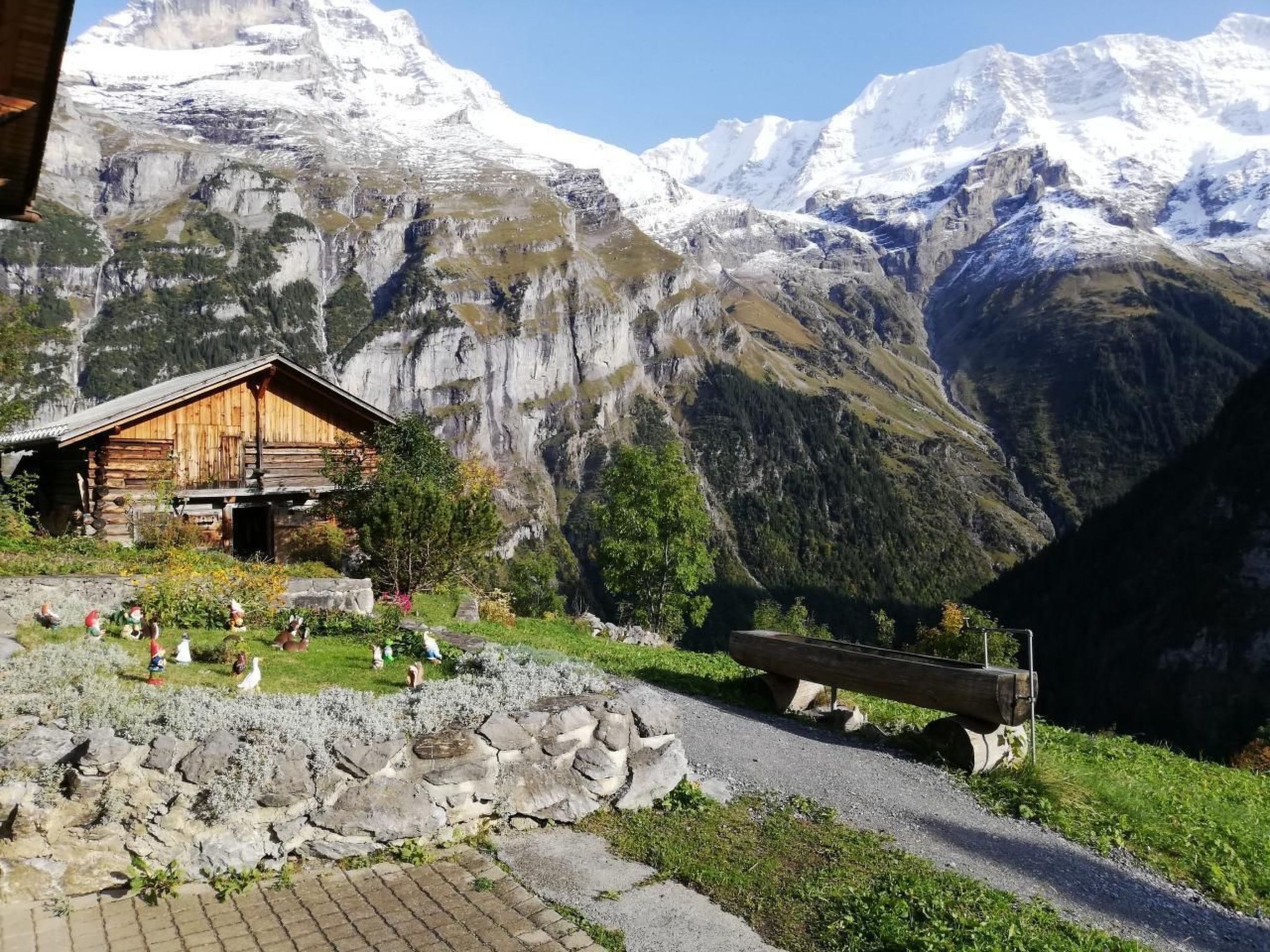 Un chalet rustique en bois, avec un sentier de pierres menant à l’entrée, entouré d’un paysage montagneux aux sommets enneigés.
