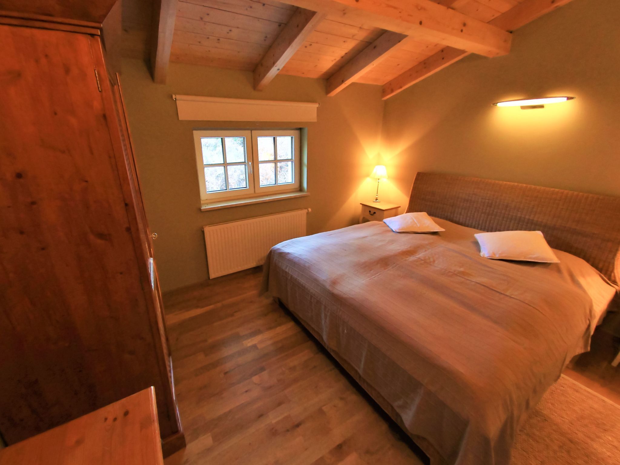 Ein Schlafzimmer mit Holzboden, einem Bett mit Kissen, einem Nachttisch mit Lampe und einem Fenster mit Jalousien.