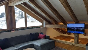 Ett mysigt vardagsrum med en hörnsoffa, en liten fåtölj i rotting och en platt-TV som är monterad på en träbalk.