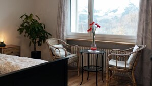 Ein Schlafzimmer mit einem Bett, einem Topf mit einer Pflanze, zwei Korbstühlen, einem kleinen runden Tisch und einer Vase mit Blumen.