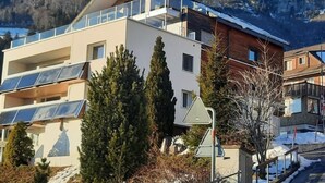 Ein modernes mehrgeschossiges Gebäude mit Balkonen, Solaranlagen und Blick auf schneebedeckte Berge.