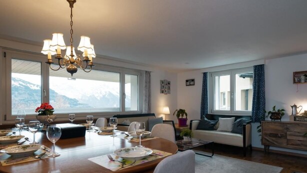 Ein Essbereich mit einem Holztisch, gedeckt für vier Personen, einer Chandelierleuchte und Blick auf Berge durch große Fenster.
