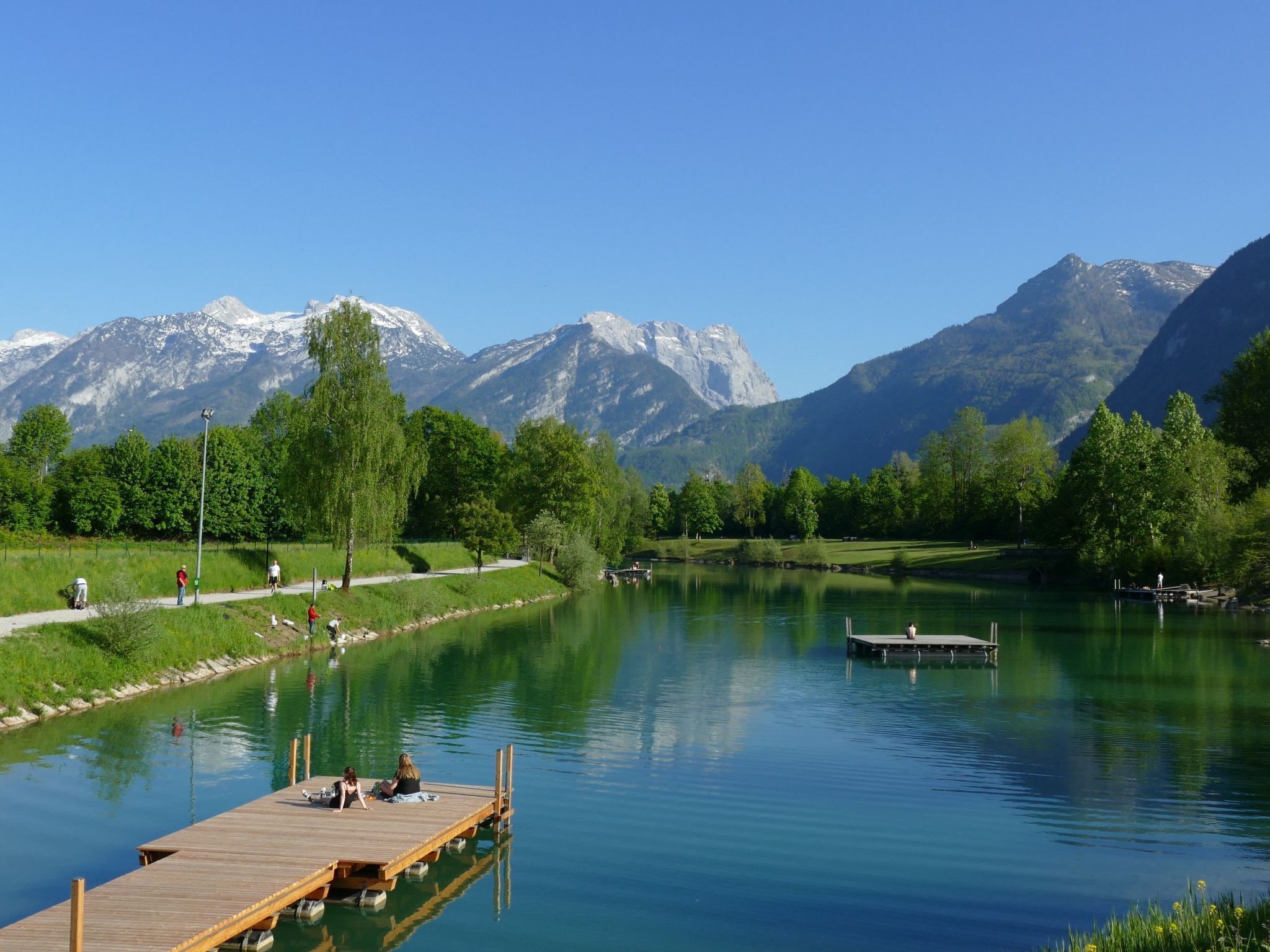 A serene lakeside scene with a wooden dock extending into the water, surrounded by lush greenery and mountains in the background.