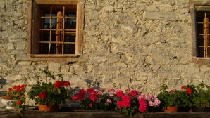 A stone building with two windows, each with wooden frames and bars. There are flower pots with red and pink flowers in front of the building. - in Sankt Koloman mit Bergblick by Interhome (St. Koloman)