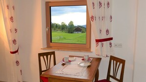 A dining area with a wooden table, two chairs, a window with a view of a green field and a building, and white curtains with red heart patterns. - in Sankt Koloman mit Bergblick by Interhome (St. Koloman)