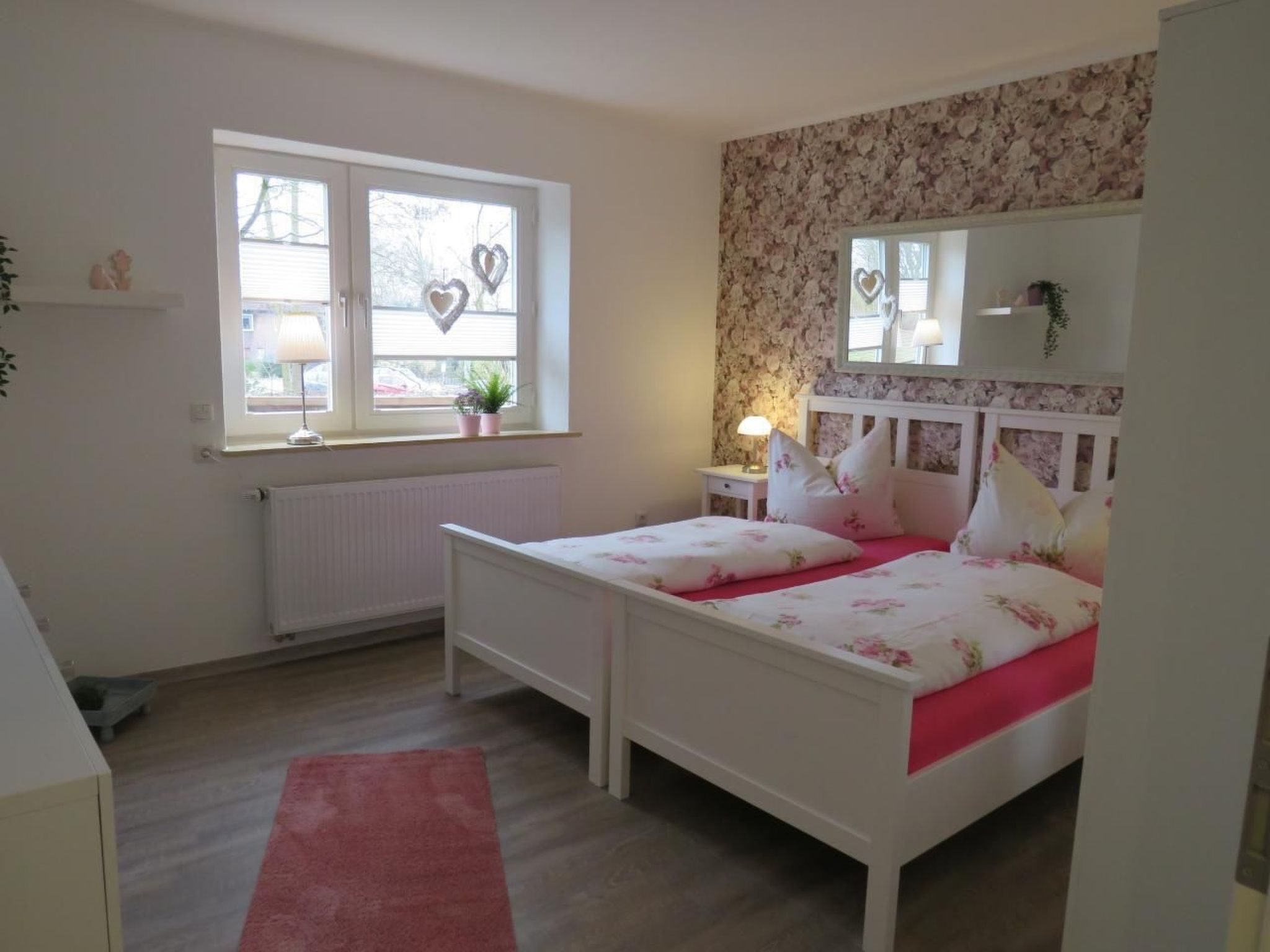 A bedroom with a bed, a window with heart-shaped decorations, a radiator, a mirror, and a pink rug.