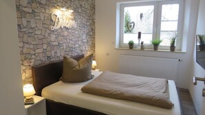 A bedroom with a stone wall, a bed with a beige blanket, two pillows, a nightstand with a lamp, and a window with plants. - Ganze Wohnung in Voslapp by Interhome (Wilhelmshaven)