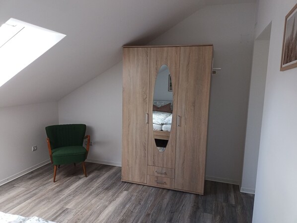 Una habitación con techo inclinado, un armario de madera con espejo, una silla verde y una cama con ropa de cama estampada.