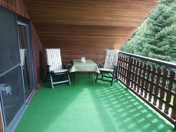 Un balcon avec une table et deux chaises, une balustrade en bois et une vue sur la verdure.