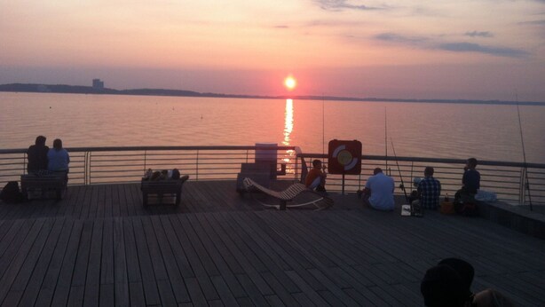 A wooden deck with people sitting and relaxing, overlooking a calm body of water during sunset. - Ostsee Perle by Interhome (Niendorf)