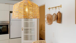 Uma cozinha com armários brancos, uma luminária pendente de fibra natural e uma porta que dá para um quarto com uma cama.