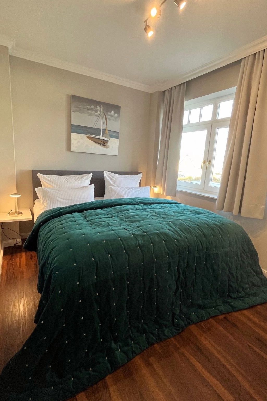 Una cama bien hecha con colcha verde, una mesita de noche con lámpara, un cuadro de un velero en la pared y una ventana con cortinas.
