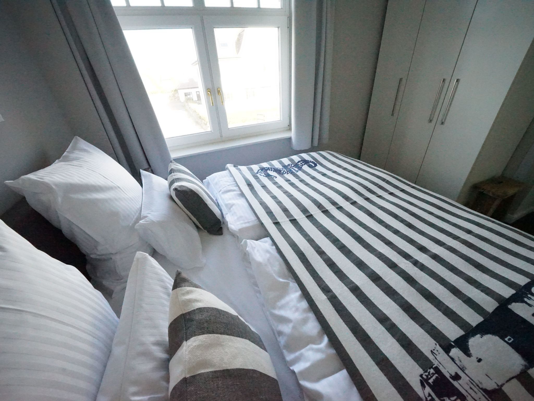 Quarto com cama, travesseiros, manta listrada e janela com cortinas.