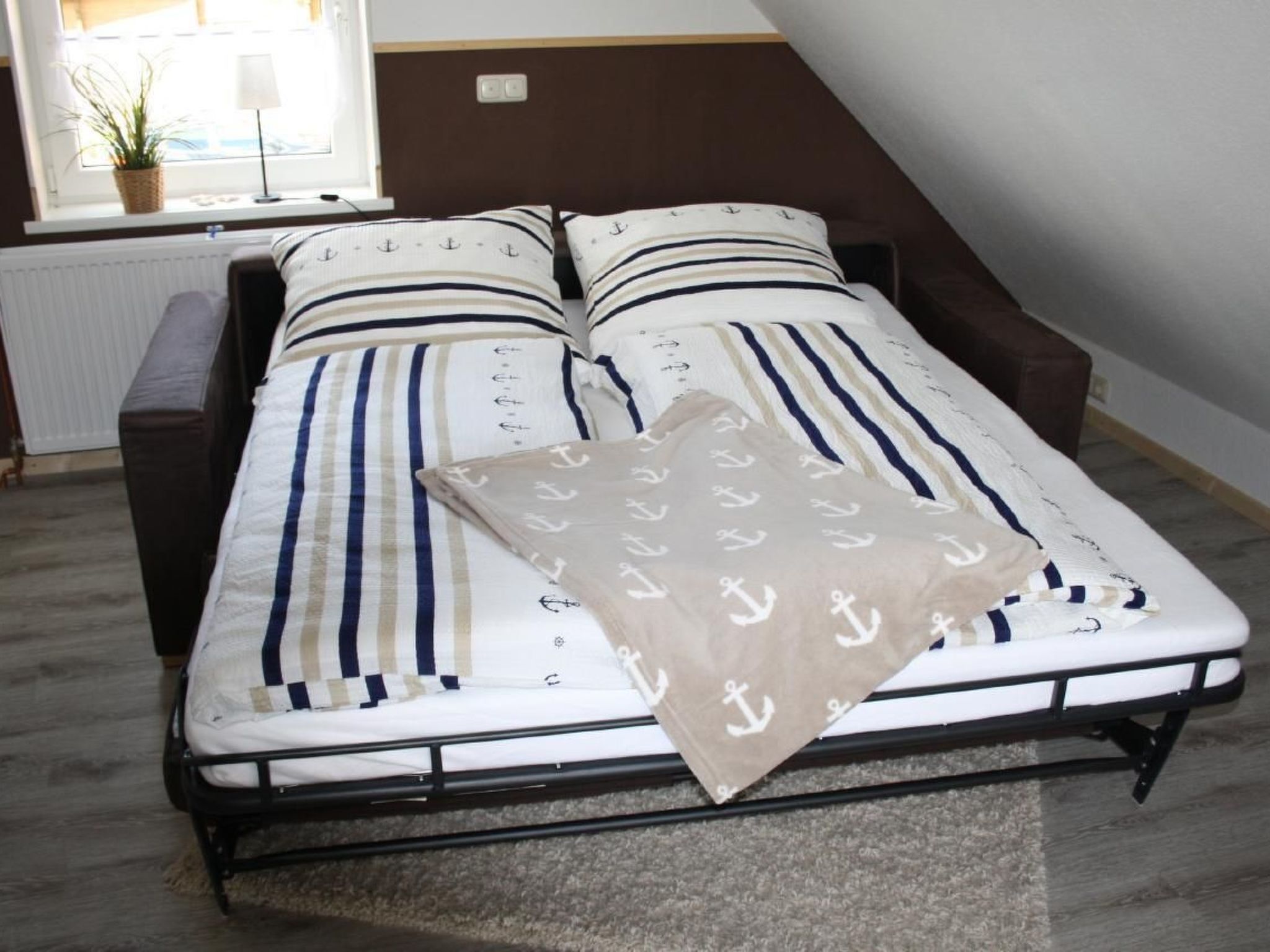 Een bed met gestreept beddengoed en een anker dessin, een nachtkastje, een lamp, een raam met een plant, en een radiator.
