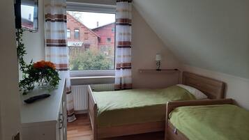 Ett litet, ombonat rum med två sängar i trä, ett element, ett fönster med randiga gardiner, en liten hylla med en lampa och en växt med orange blommor.