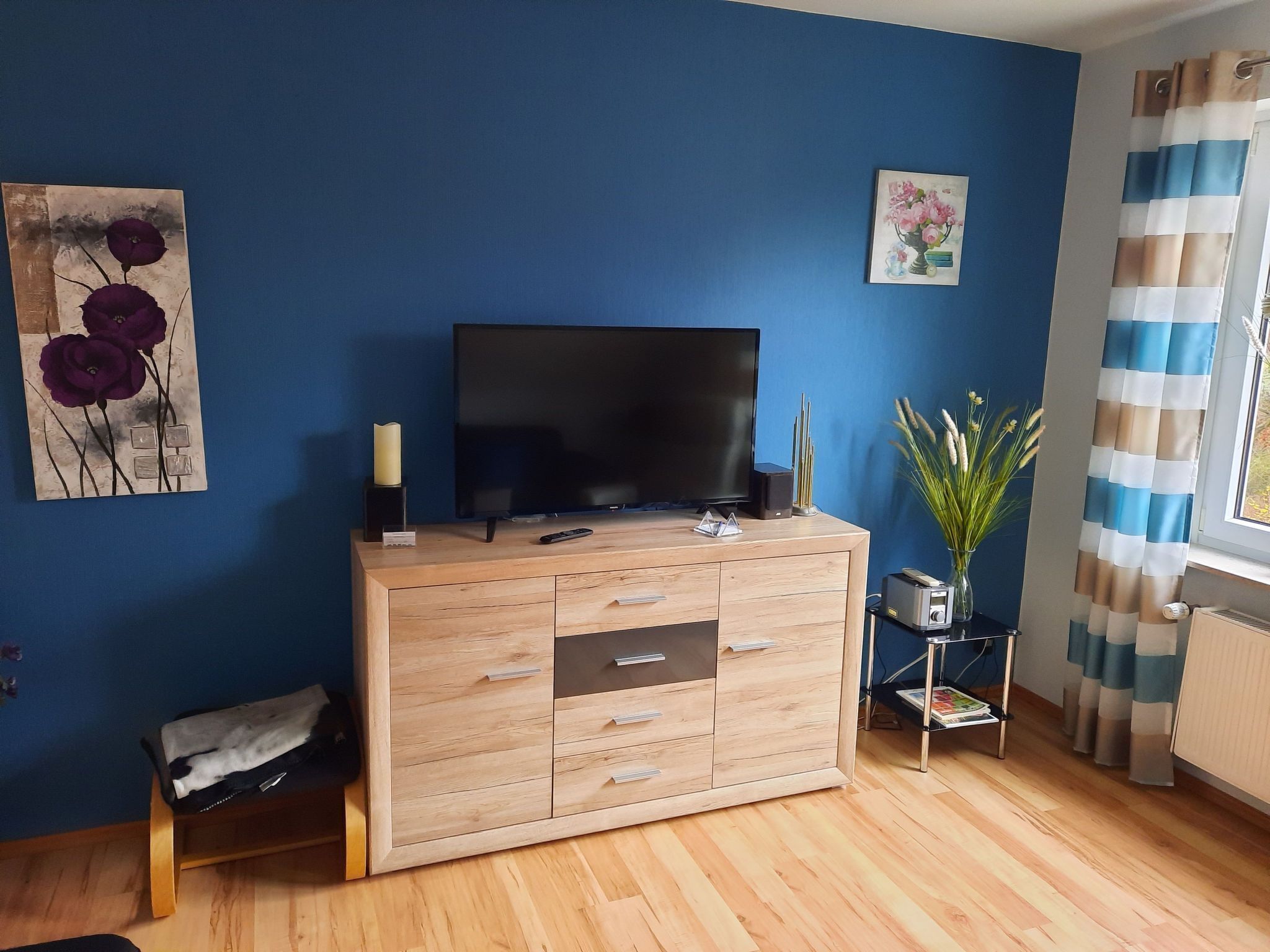 Sala de estar com um armário de madeira, uma TV de tela plana, uma mesinha lateral com um vaso e uma janela com cortinas.