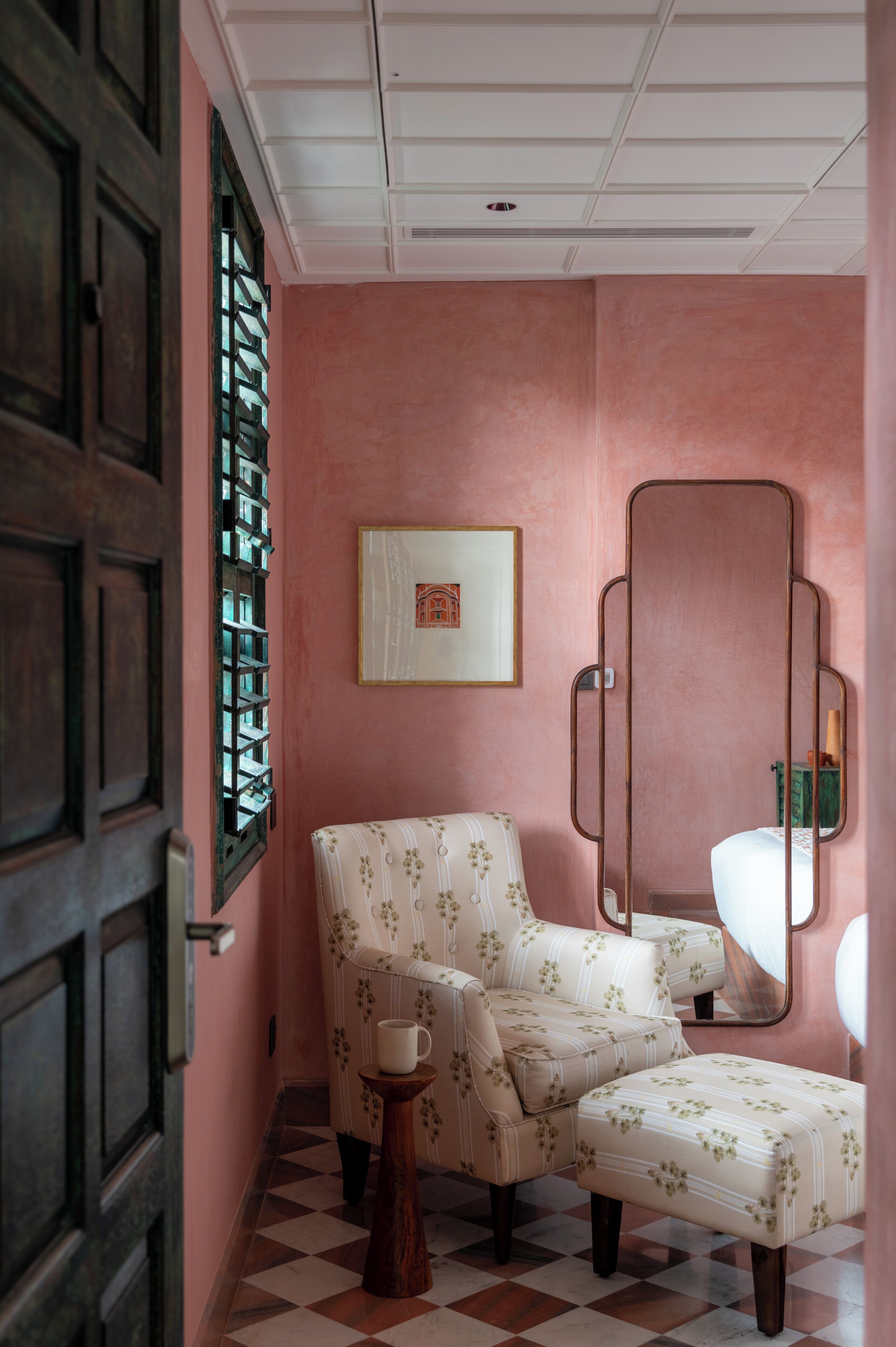 A room with a checkered floor, a floral upholstered chair, a small wooden stool, a framed picture on the wall, and a large mirror.
