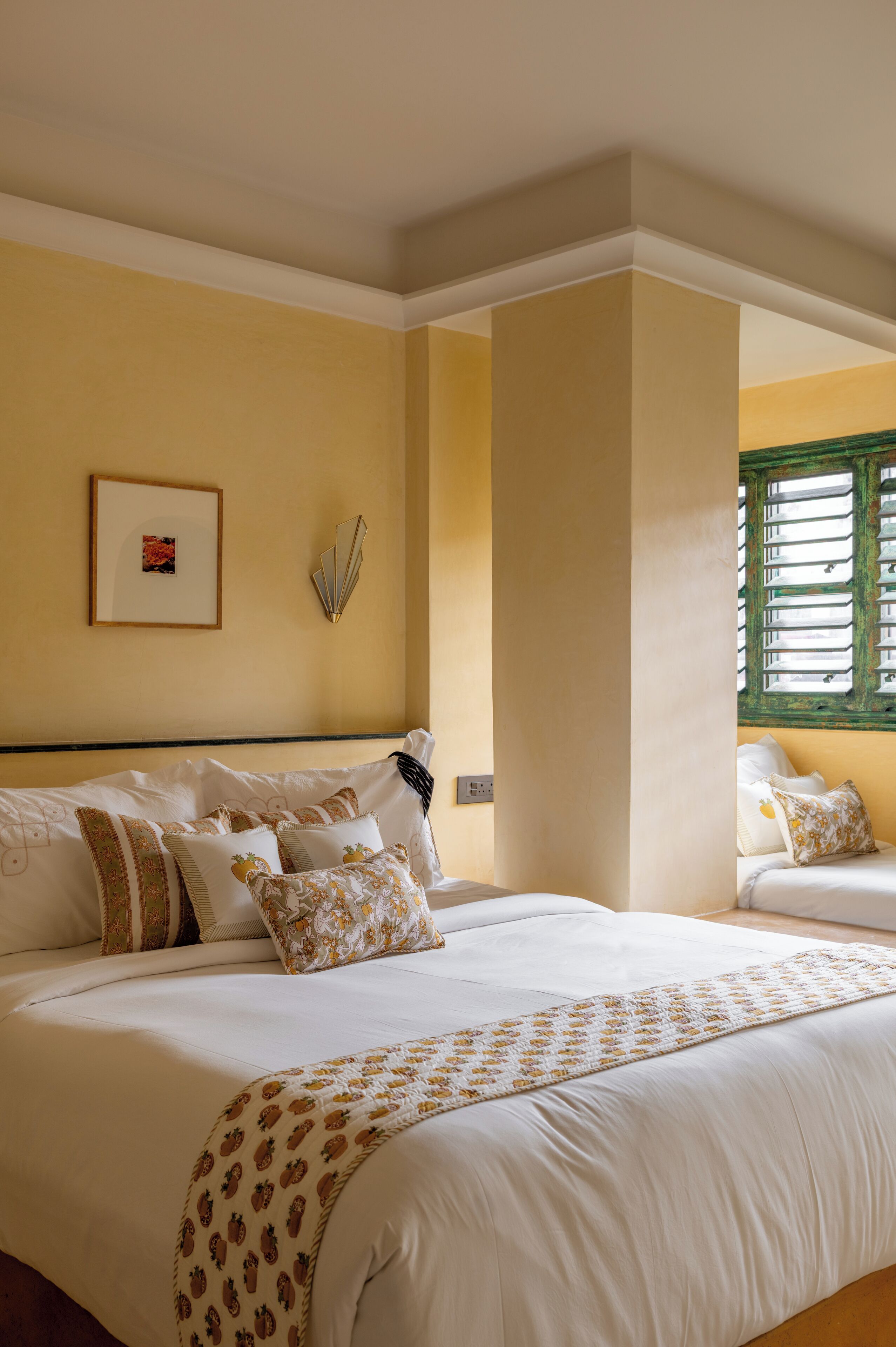 A neatly made bed with a patterned bedspread and multiple pillows, a framed picture on the wall, and a window with blinds.