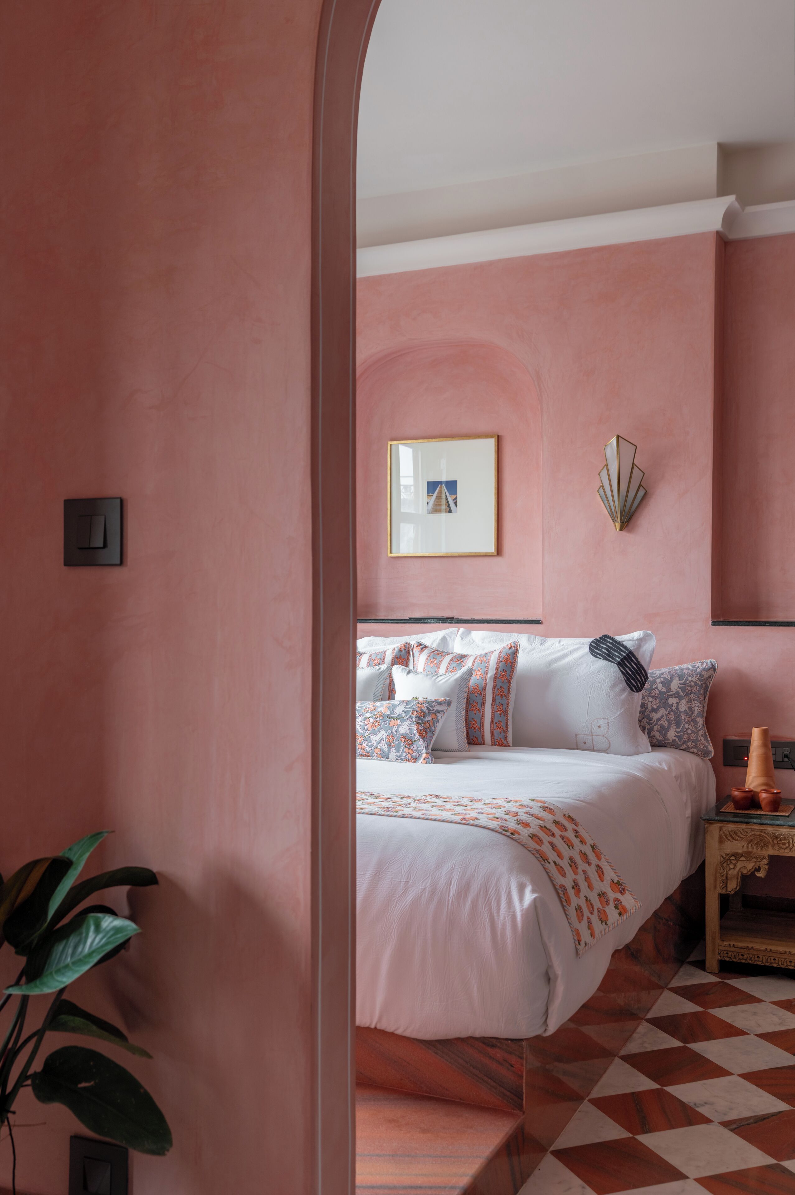 A bedroom with a checkered floor, a bed with patterned pillows, a wall-mounted light fixture, and a framed picture on the wall.