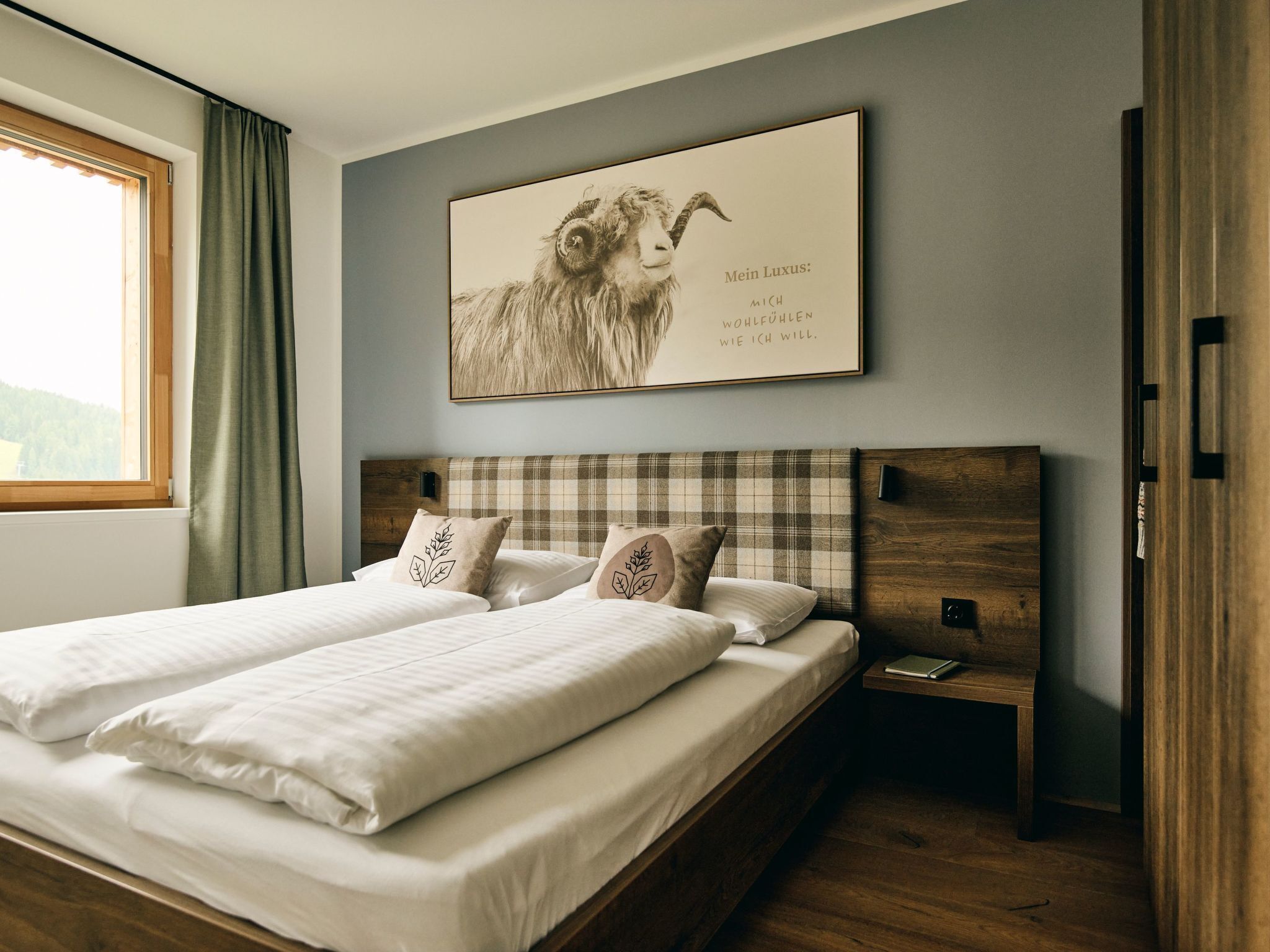 A double bed with a wooden headboard, two pillows with leaf designs, a bedside table with a book, and a framed picture on the wall.