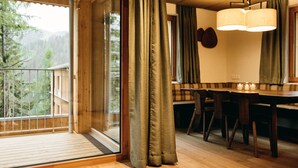 Interior de madera con zona de comedor, un ventanal con cortinas y vistas a un área boscosa.