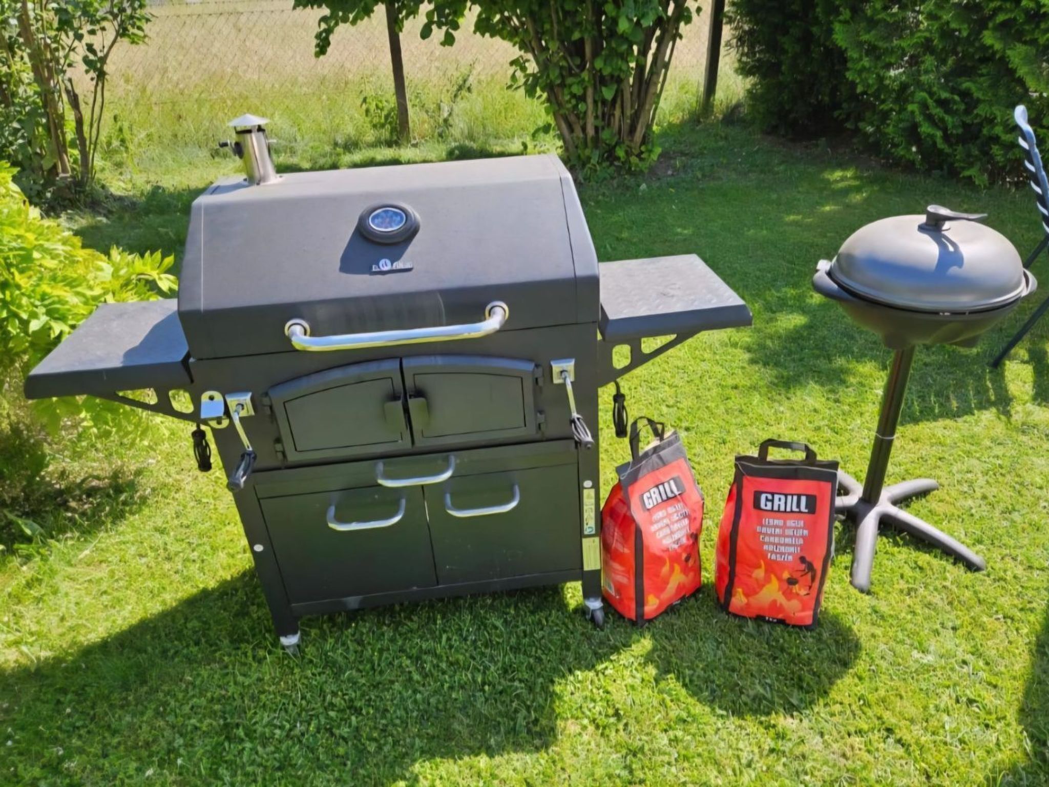 Un barbecue avec une table d’appoint et deux sacs étiquetés « BARBECUE », le tout installé dans un espace extérieur verdoyant.