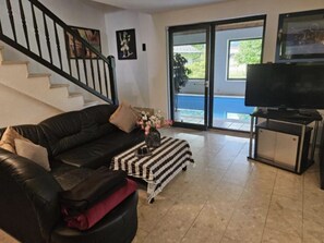 Una sala de estar con un sofá de cuero negro, un pequeño reposapiés, un televisor sobre un mueble bajo y una escalera.