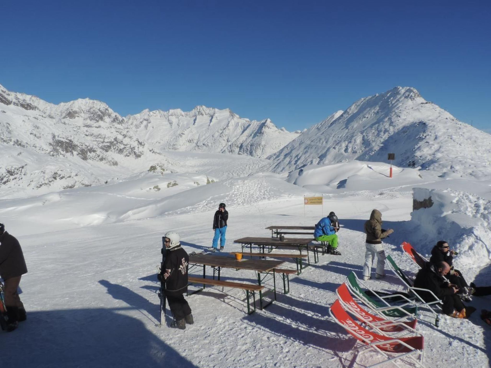 Escena nevada de muntanya amb cel serè, que mostra una zona de pícnic amb taules i bancs, i una rampa per fer snowboard.