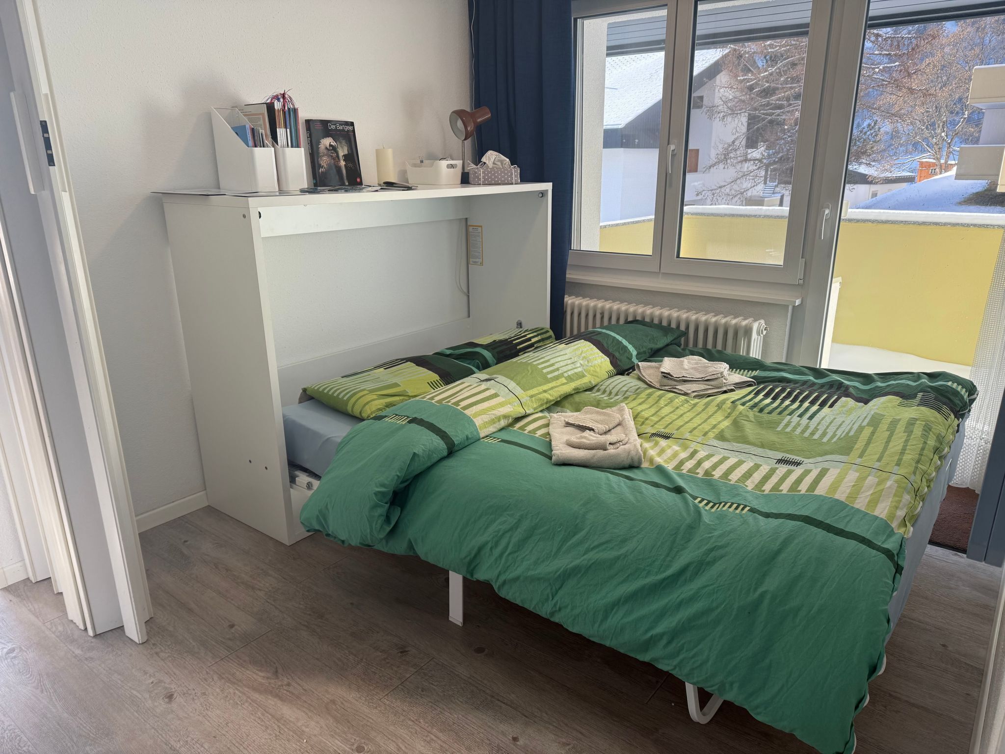 A single-bed room with a green bedspread, a white dresser, a window with a view of a snowy landscape, and a radiator under the window.