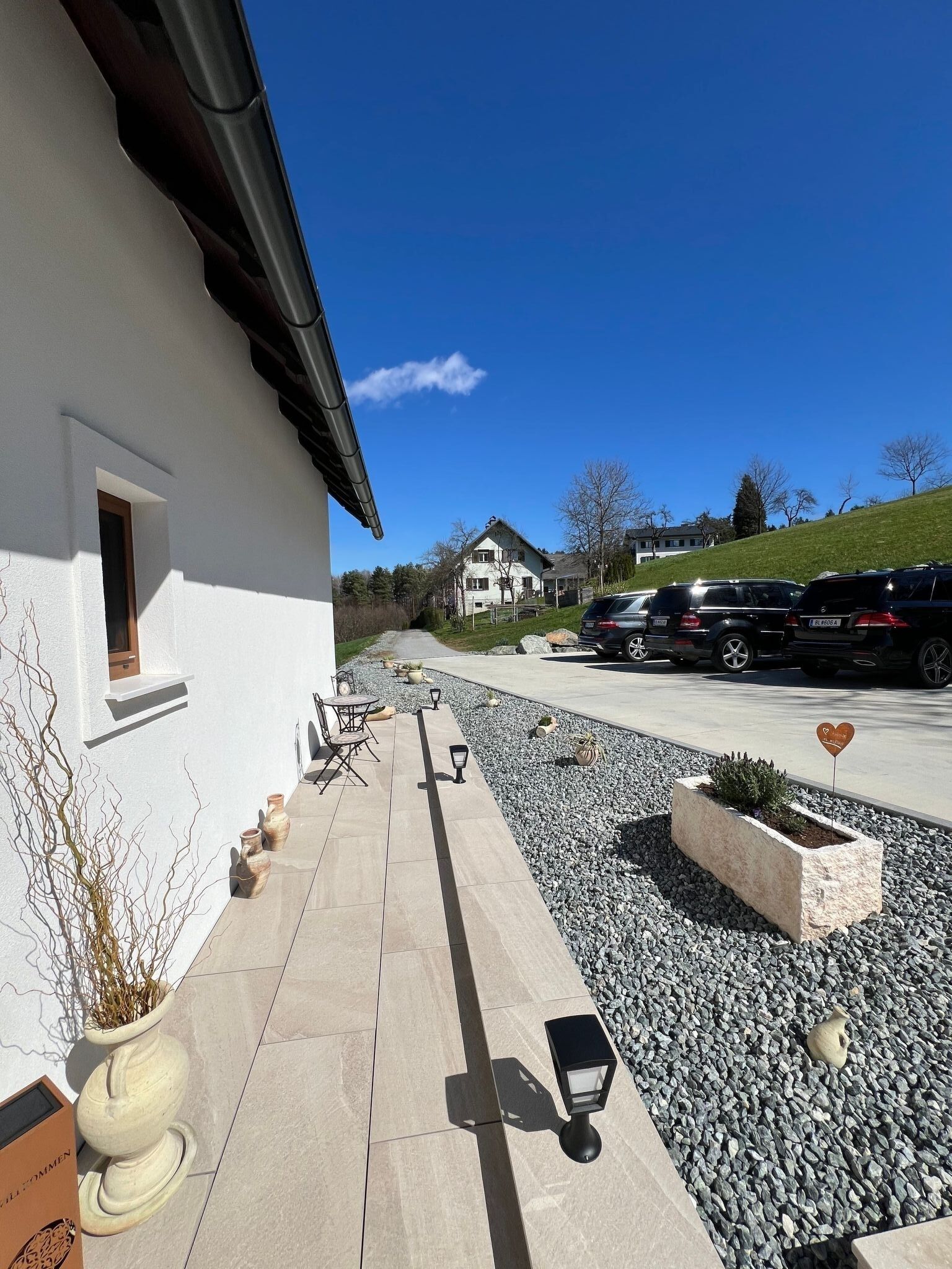 Un bâtiment à l’extérieur blanc, avec un chemin pavé, des places assises extérieures et un espace de stationnement où sont garées des voitures.