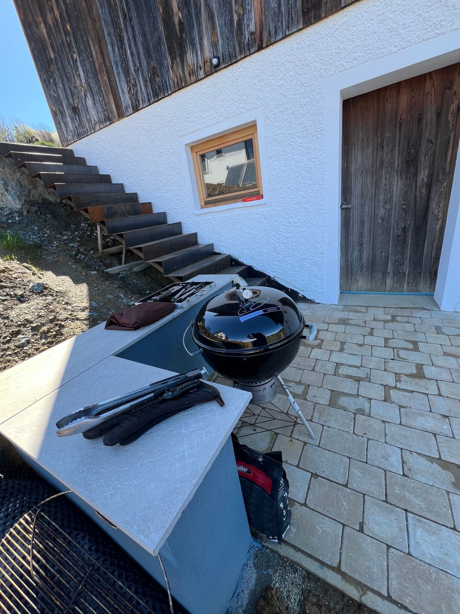 En grillopsætning på en terrasse med en hvid væg og trætrapper, der fører op til en dør.