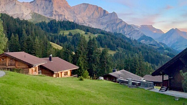 Un chalet de montagne doté d’un toit en pente, entouré de collines verdoyantes et avec, en toile de fond, des montagnes escarpées.