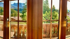 Une vue panoramique à travers une fenêtre à cadre en bois, dotée d’une poignée en métal, donnant sur un paysage montagneux parsemé d’arbres et sur un bâtiment.