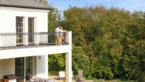 A two-story house with a balcony, outdoor seating area, and a view of a dense forest.