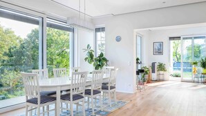 A modern dining area with a white table and chairs, large windows, and a view of greenery outside.