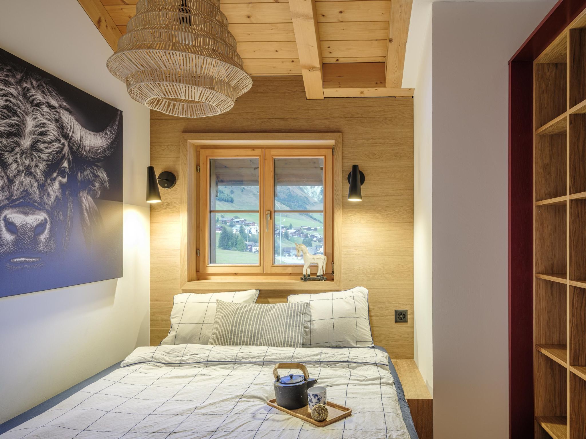A bedroom with a bed, a window with a view of a village, a wooden ceiling, and a wall-mounted lamp.
