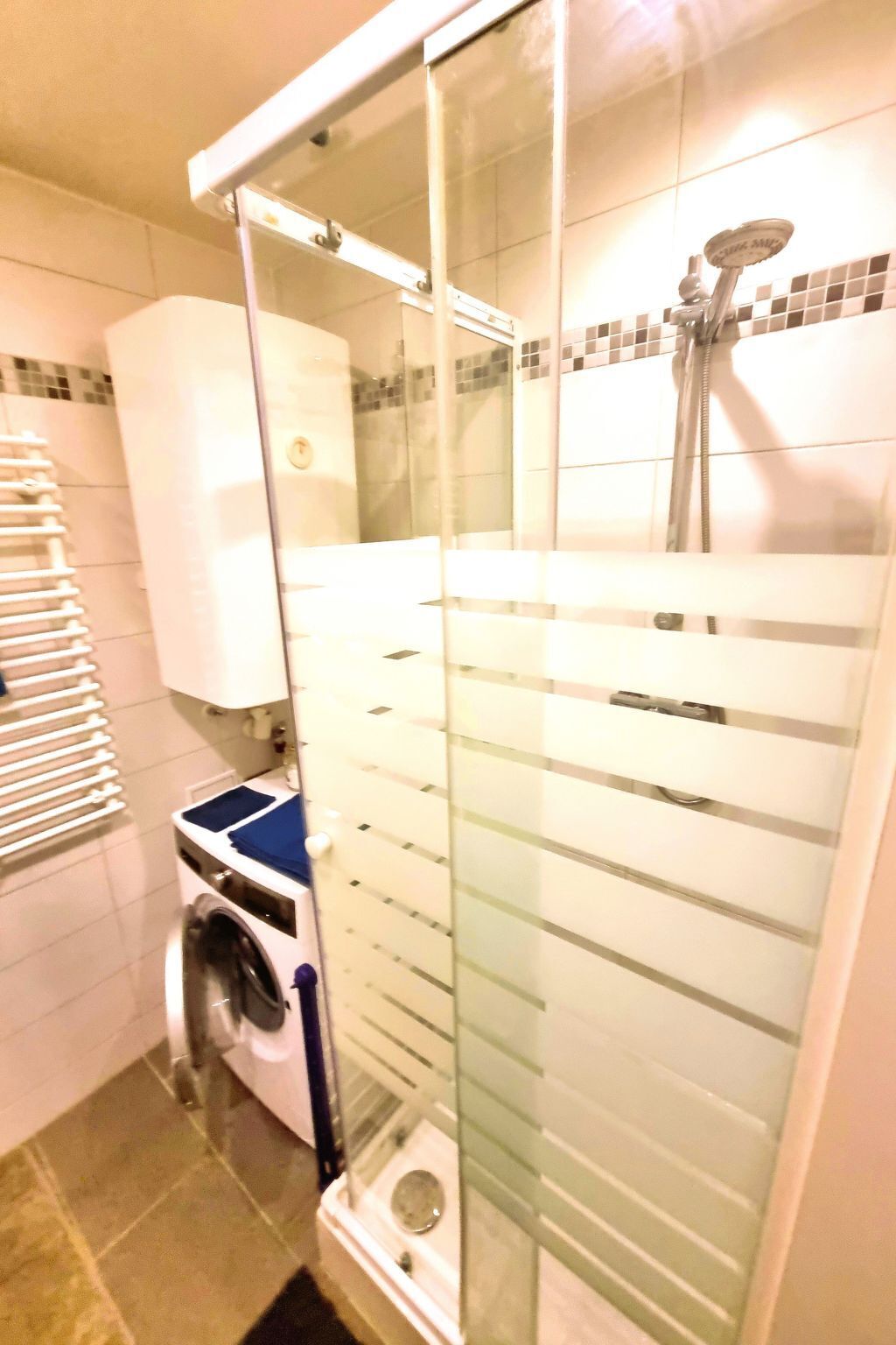 A compact bathroom with a glass shower enclosure, a built-in washing machine, and a tiled floor.