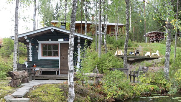 A cabin with a deck and a small dock extending into a pond, surrounded by trees.