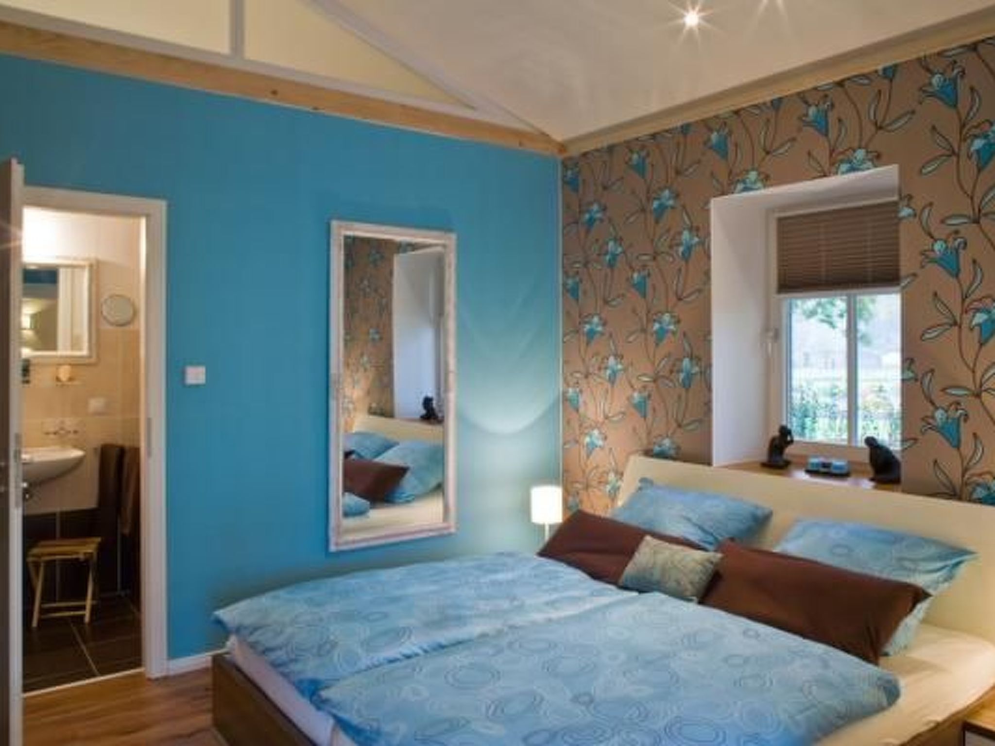 A bedroom with a bed, a mirror, a window with blinds, and a bathroom visible through an open door.