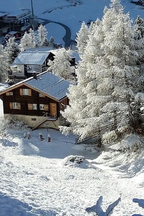 Una cabaña de madera con techo inclinado, rodeada de árboles cubiertos de nieve y un telesilla al fondo.