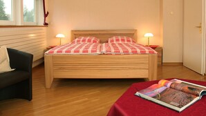 Ein ordentlich eingerichtetes Schlafzimmer mit einem hölzernen Bett, zwei Nachttischlampen, einem schwarzen Sessel und einer rotumschlagenen Zeitschrift auf einem kleinen Tisch.