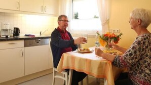 Ein Essbereich mit Tisch für zwei Personen, einem mit Vorhängen versehenen Fenster und einer Küche im Hintergrund.