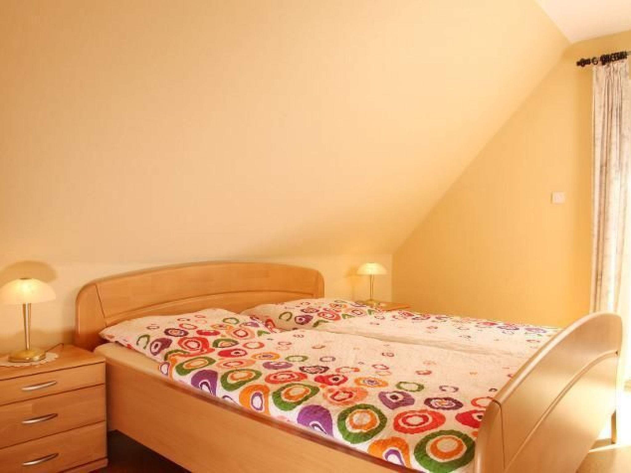 Una cama individual con una colcha estampada de colores en una habitación con techo inclinado y ventana con cortinas.