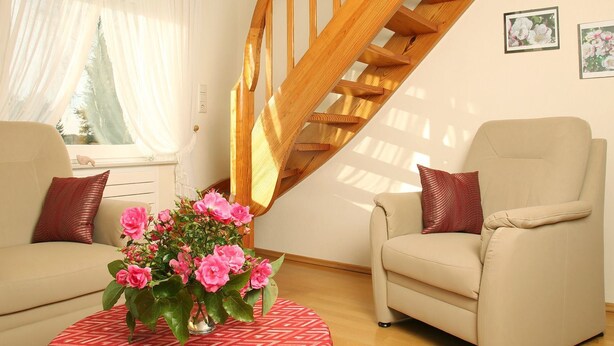 Ein Wohnzimmer mit einer Holztreppe, zwei beige Armlehnstühlen, einem rot gemusterten Hocker und einer Vase mit rosa Blumen.