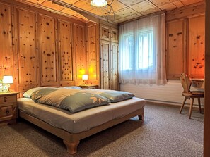 A bedroom with a bed, bedside tables, a chair, and a window with curtains. - Apartment Cervin by Interhome (Zermatt)