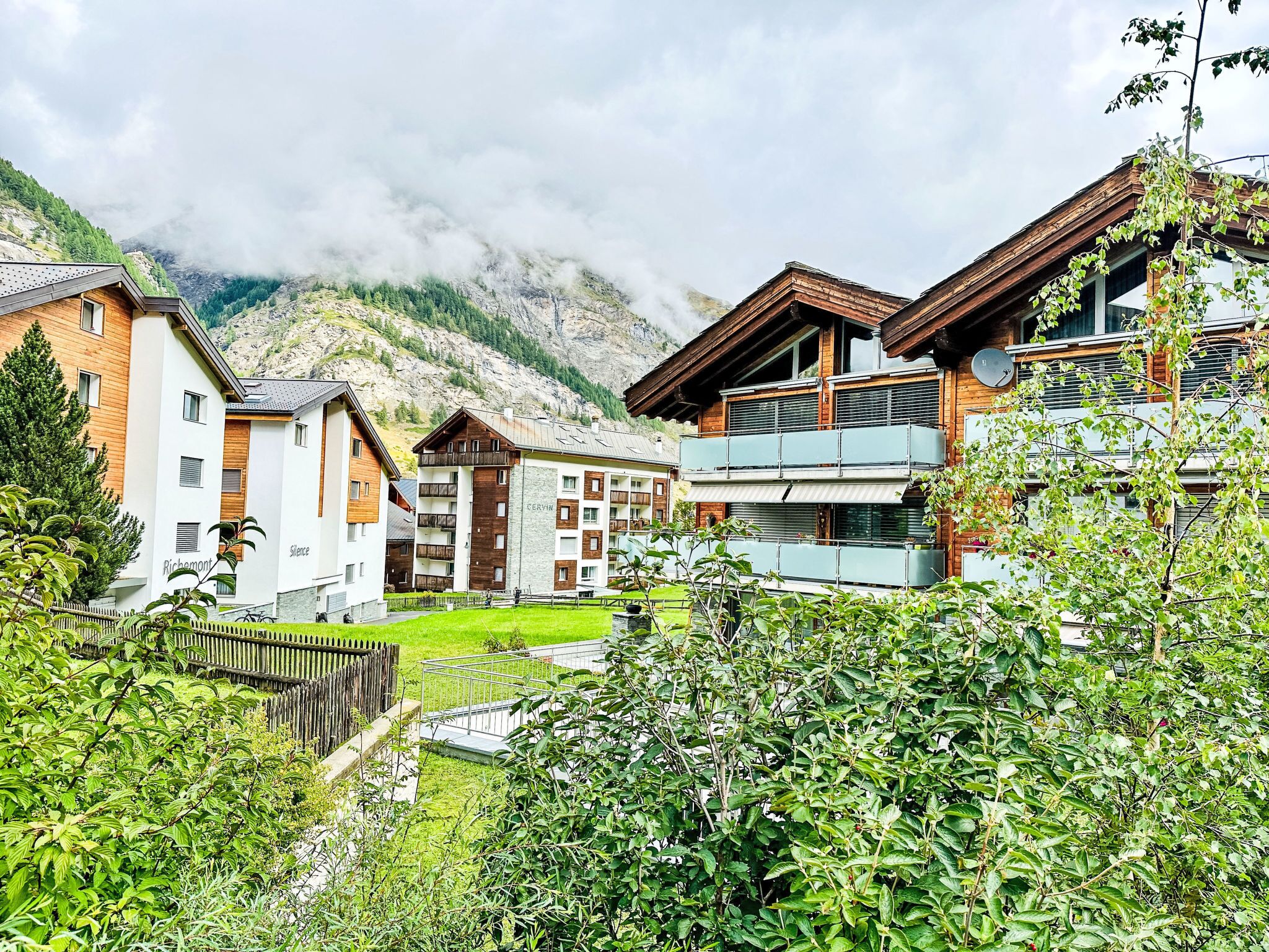 Ein Bergdorf mit Holzhäusern, einem grünen Innenhof und Blick auf einen Berg.