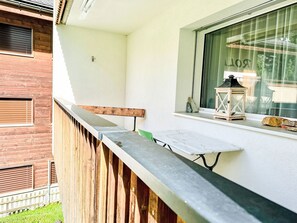 Uma varanda com guarda-corpo de madeira, uma mesa pequena, uma cadeira verde e uma janela com vista para um edifício.