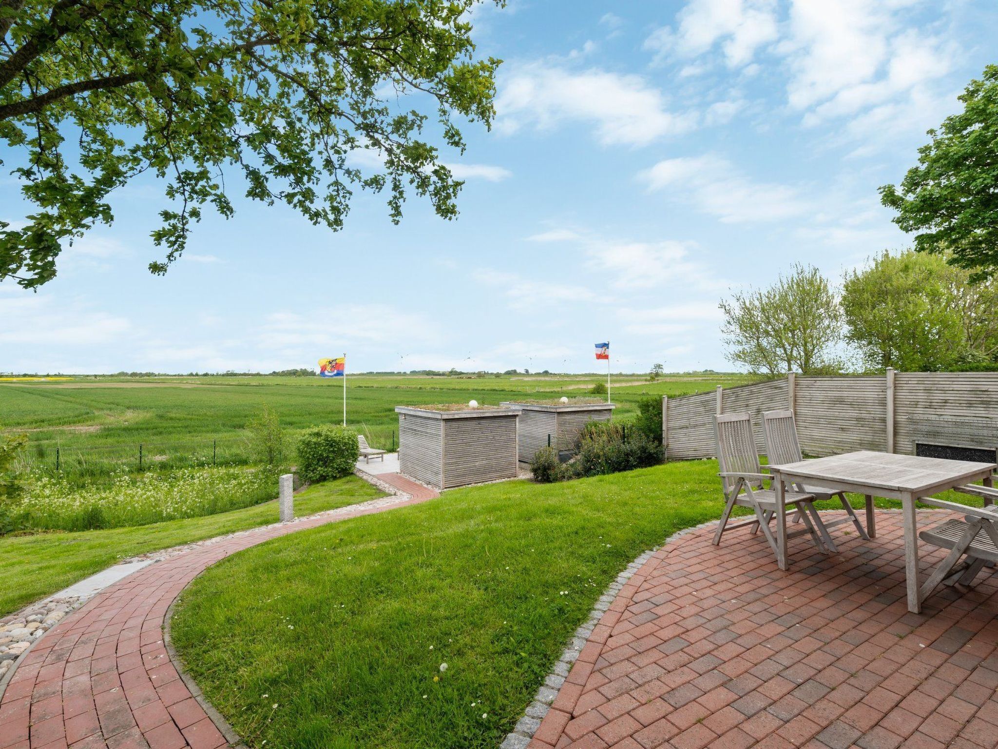 A patio with a brick pathway, wooden furniture, and flags in a rural setting.